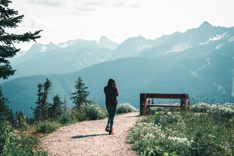 Eine Frau läuft einen Wanderweg entlang, der zu einem Aussichtspunkt mit Bank führt, von welchem aus sich ein spektakulärer Blick auf die kanadischen Rocky Mountains bei Revelstoke bietet