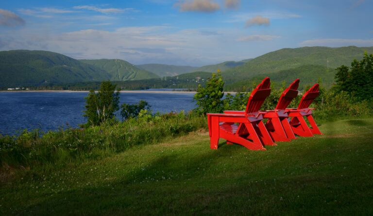 Drei rote Adirondack Chairs stehen in einer Reihe am grünen Ufer des St.-Lorenz-Golfes bei Ingonish