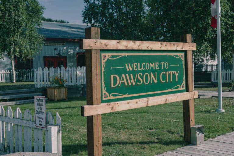 Willkommensschild für Besucher von Dawson City, umgeben von weißen Zäunen um eine kleine Grünfläche mit dem historischen Feuerwehrgebäude im Hintergrund