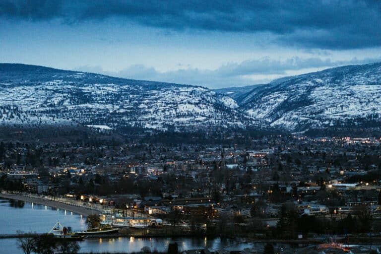 Lichter der Stadt Penticton bei Einbruch der Nacht mit Blick auf das Ufer des Okanagan Lake mit Anlegestelle in Vordergrund und den schneebedeckten Bergen im Hintergrund