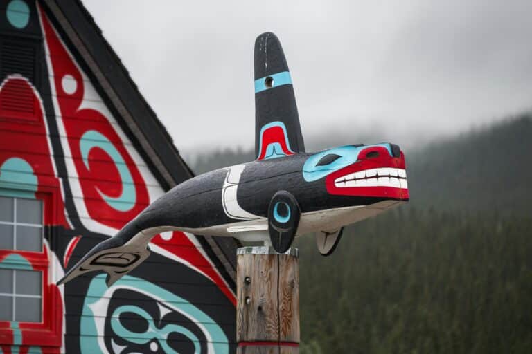 Totem-Schnitzerei in Carcross in Form eines Wals in den Farben schwarz, blau, weiß und rot, vor einem Gebäude stehend, das mit Mustern in den selben Farben bemalt ist